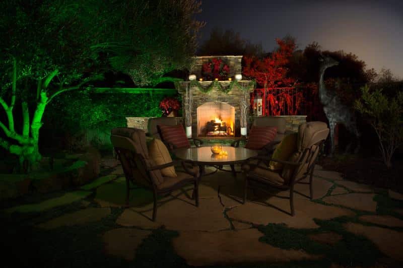 Holiday color landscape lighting illuminating a backyard patio and outdoor fireplace